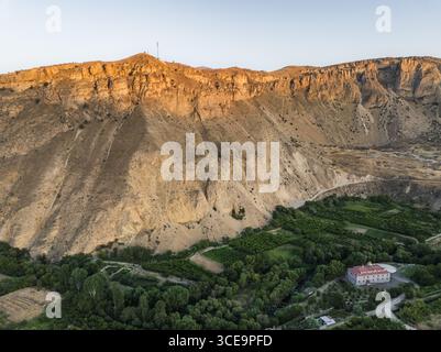 Vue aérienne des falaises escarpées et embrassées par le soleil se dessinent au-dessus de la vallée verdoyante parsemée d'un bâtiment solitaire, projetant de longues ombres dans la lumière dorée, Areni, province de Vayots Dzor, Arménie. Banque D'Images