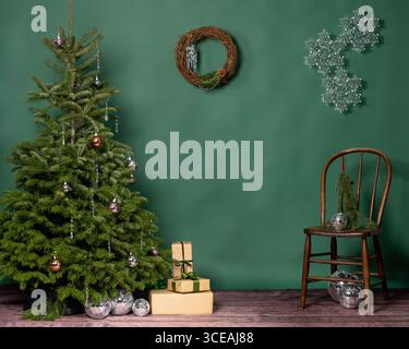 Scène de Noël minimaliste avec sapin décoré, chaise en bois vintage, cadeaux emballés et ornements de vacances sur fond vert. Banque D'Images