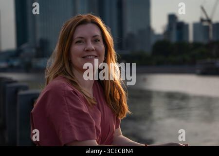 Femme avec de longs cheveux souriant dans la lumière chaude du coucher du soleil, assise près de la rivière avec des gratte-ciel modernes floues en arrière-plan Banque D'Images