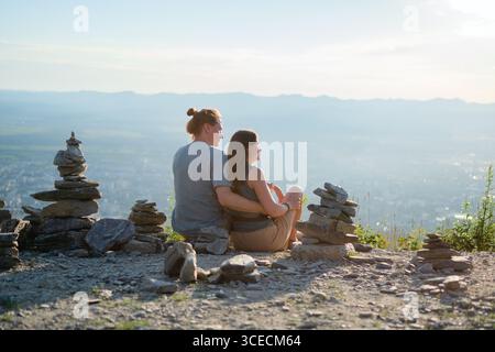 Jeune couple assis dans les bras par derrière tout en admirant un panorama urbain à couper le souffle lors de leur randonnée. Idéal pour les thèmes romantiques et d'aventure. Banque D'Images