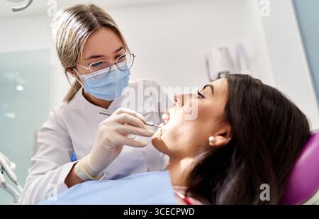 Un dentiste portant un masque et des gants examine les dents d'un patient, à l'aide d'outils dans une clinique dentaire moderne. Le patient est confortablement incliné dans la dent Banque D'Images