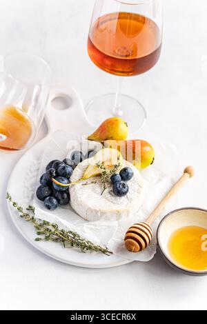 Plateau de fromage élégant avec brie crémeux, poires mûres et myrtilles juteuses, accompagné d'un verre de vin ambré sur une table blanche, offrant un sophisti Banque D'Images