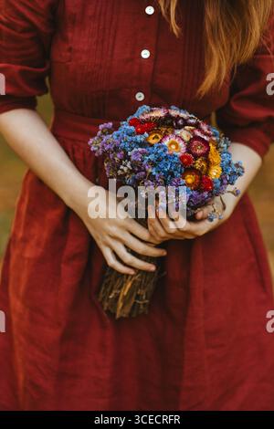 Une femme dans une robe rouge tient doucement un bouquet vibrant de fleurs séchées au Québec, Canada. Les couleurs riches et les tons chauds capturent parfaitement l'essence Banque D'Images