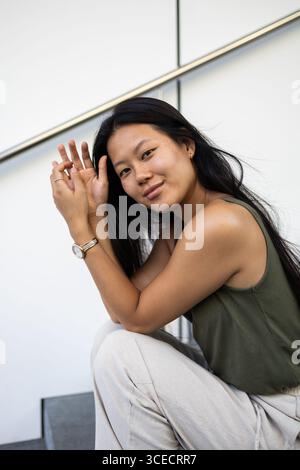 Une femme chinoise est assise à l'extérieur sur les marches de la ville, profitant d'une journée d'été lumineuse. Elle semble détendue et contente, avec de longs cheveux qui descendent en cascade le long de son épaule Banque D'Images