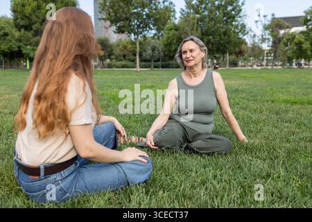 Une mère et sa fille partagent un moment paisible assis sur l'herbe dans un parc ensoleillé, profitant de la compagnie et de l'air frais de l'extérieur. C'est un jour de Banque D'Images