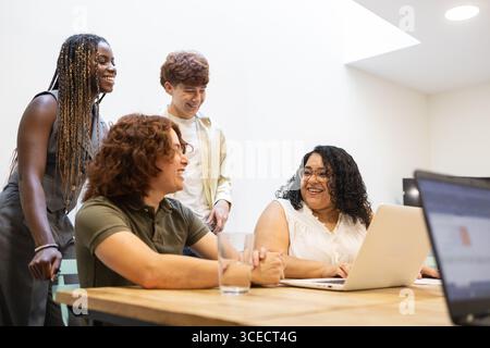 Un groupe diversifié de collègues collaborent dans un environnement de bureau moderne. Ils sont engagés dans une discussion positive autour d'une table avec des ordinateurs portables, en encourageant Banque D'Images