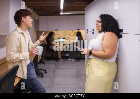 Vue latérale de deux jeunes professionnels collaborant dans un espace de coworking moderne. Deux personnes sont engagées dans la conversation tandis que d'autres travaillent sur des ordinateurs portables Banque D'Images