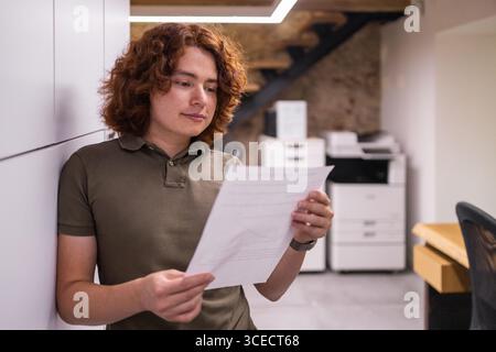 Un jeune homme aux cheveux bouclés s'appuie contre un mur tout en lisant un document dans un espace de coworking confortable et bien éclairé pendant l'été. Banque D'Images