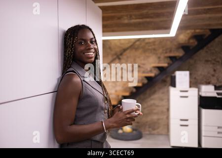 Jeune femme africaine aux tresses se tient souriante tenant une tasse, profitant d’une pause dans un espace de coworking contemporain. Le cadre est moderne avec un ton chaleureux Banque D'Images