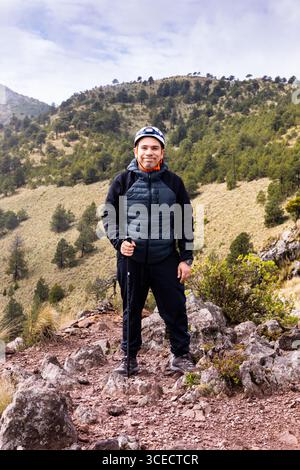 Un randonneur portant un équipement de plein air se tient en toute confiance sur un sentier de montagne rocheux, entouré d'arbres verdoyants et d'une vue lointaine sur les collines ondulantes sous un Banque D'Images