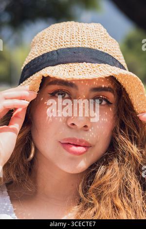 Une jeune femme latino-américaine profite d'une journée ensoleillée à l'extérieur, portant un chapeau de paille tendance. Son expression décontractée et son style décontracté incarnent un style de vie insouciant Banque D'Images