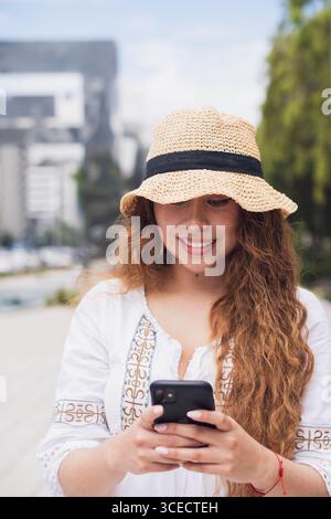 Une jeune femme latino-américaine sourit en utilisant son smartphone à l’extérieur, incarnant un style de vie moderne et détendu. Sa tenue décontractée et son chapeau se plaignent Banque D'Images