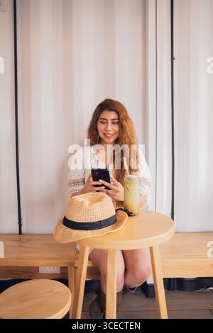 Jeune femme latino-américaine se détendant dans un café, sirotant une boisson rafraîchissante, et souriant en regardant son smartphone, un exemple de style de vie moderne Banque D'Images