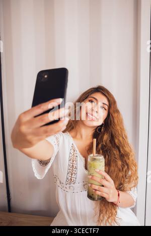 Une jeune femme latino-américaine profite de son style de vie en prenant un selfie avec son smartphone tout en tenant une boisson de thé à bulles tendance, mettant en valeur une ambiance détendue Banque D'Images