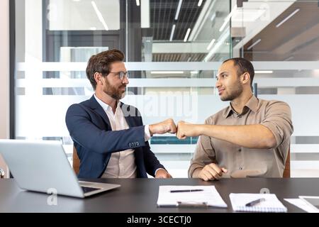 Deux collègues dans un cadre professionnel font preuve de camaraderie avec un coup de poing, symbolisant le travail d'équipe réussi et le respect mutuel, dans un bureau contemporain cloisonné de verre. Banque D'Images