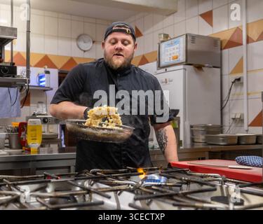 Un chef dans un uniforme noir prépare habilement un plat dans une cuisine professionnelle, mettant en valeur l'habileté et la concentration. La toile de fond de la cuisine présente variou Banque D'Images