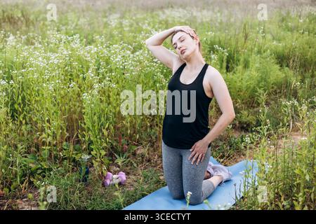 Une femme dans un débardeur noir et leggings gris effectue une pose de yoga stretch au cou sur un tapis dans un champ luxuriant, entouré de verdure et de fleurs sauvages, enjo Banque D'Images
