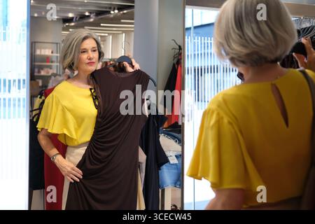 Une femme mature examine une élégante robe marron devant un miroir à l'intérieur d'un magasin de vêtements. Son expression réfléchie suggère qu'elle envisage de le faire Banque D'Images