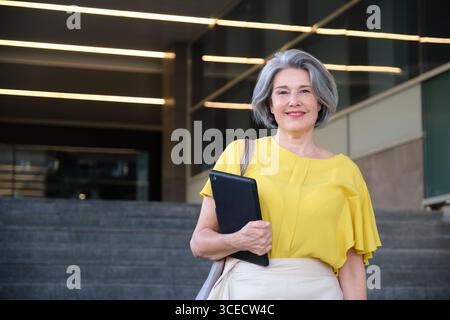 Une femme mature confiante dans une blouse jaune se tient à l'extérieur d'un bâtiment moderne, tenant une tablette et souriant chaleureusement, incarnant le professionnalisme et la joie i. Banque D'Images
