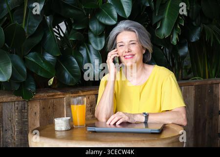 Femme mûre, souriante dans une blouse jaune, aime un appel téléphonique tout en étant assise à une table en bois avec un ordinateur portable et un verre de jus d'orange, entouré par Banque D'Images