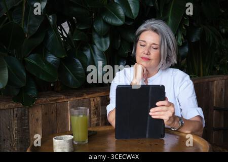 Une femme mature aux cheveux gris est assise à une table de café en bois, lisant sur sa tablette. Entourée de verdure luxuriante, elle profite de son temps avec une boisson verte Banque D'Images