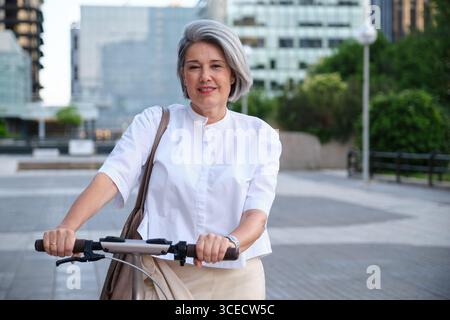 Une femme mature avec les cheveux gris courts chevauchant un scooter électrique dans un cadre urbain. Elle porte une blouse blanche et souriante, transmettant un sens de fr Banque D'Images