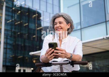 Femme mature souriante tout en tenant un smartphone à l'extérieur, vêtue d'une chemise blanche décontractée, profitant du temps libre dans un cadre urbain avec du verre moderne Banque D'Images