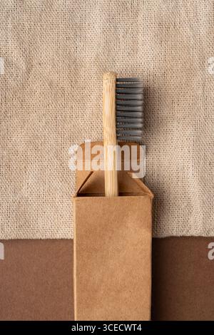 Vue de dessus d'une brosse à dents en bambou partiellement dans une boîte en carton brun, placée sur un fond textile beige texturé. Mettant l'accent sur l'environnement et zéro Banque D'Images