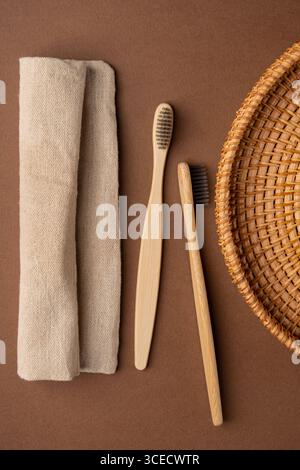Vue de dessus de deux brosses à dents en bambou à côté d'une serviette en lin beige et d'un panier en osier, placés sur un fond de tissu marron, mettant en valeur l'environnement Banque D'Images
