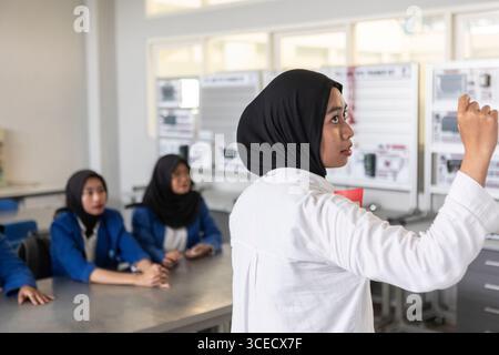 Un enseignant asiatique musulman, portant un hijab, pointe vers le tableau tout en discutant avec des étudiants enthousiastes. La scène capture un l diversifié et interactif Banque D'Images
