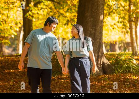 Un joyeux couple asiatique marche main dans la main à travers un parc ensoleillé couvert de feuilles d'automne. Le cadre serein capture un moment de joie et de compagnie Banque D'Images
