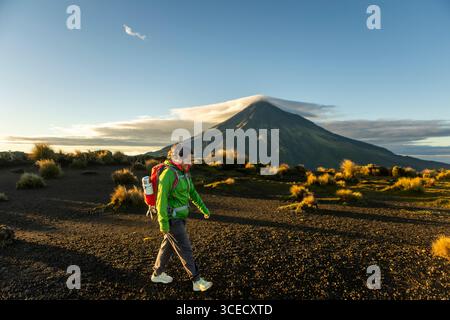 Une femme randonneuse marche à travers le terrain accidenté près du mont Taranaki, en Nouvelle-Zélande, alors que le soleil se lève, projetant une lueur dorée sur le paysage volcanique. PE Banque D'Images