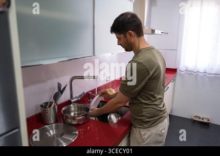 Un homme dans une chemise verte lave la vaisselle dans une cuisine moderne avec des comptoirs rouges. La scène met en évidence la corvée quotidienne de maintenir la propreté de la cuisine Banque D'Images