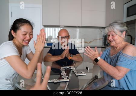 Une joyeuse famille multigénérationnelle jouant des dominos autour d'une table de cuisine. Trois membres de la famille sourient et applaudissent tout en profitant de leurs loisirs Banque D'Images