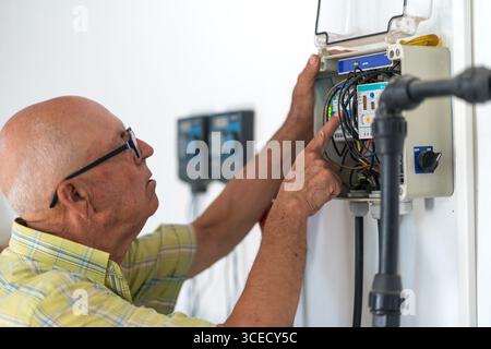 Un homme âgé portant des lunettes travaille attentivement sur un panneau électrique, ajustant les fils et utilisant des outils. Le cadre met en évidence son attention focalisée A. Banque D'Images