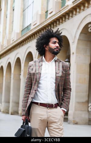 Un vendeur d'assurance mixte aux cheveux afro se tient en toute confiance à l'extérieur, vêtu d'un blazer à carreaux et d'une chemise blanche. L'arrière-plan présente classique Banque D'Images