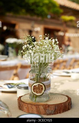 Une pièce maîtresse de table de mariage rustique avec de délicates fleurs blanches dans un pot maçon orné de dentelle, placé sur une dalle de bois. Idéal pour boho romantique Banque D'Images
