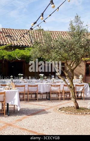 Cadre de mariage extérieur élégant présentant de longues tables ornées de draps blancs et de pièces maîtresses florales. Le fond rustique présente un charmant co Banque D'Images