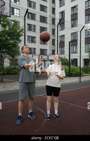 Un père et sa fille aiment jouer au basket-ball ensemble sur un terrain extérieur entouré d'immeubles d'appartements modernes. L'activité favorise le fitness et Banque D'Images