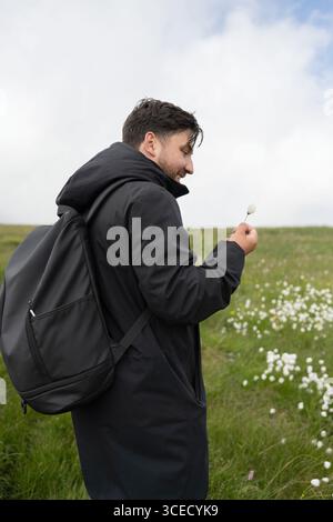 Un randonneur en veste noire se tient au milieu d'un champ vibrant de fleurs blanches sur un sentier de montagne, profitant de la beauté naturelle et de l'atmosphère sereine sous-jacente Banque D'Images