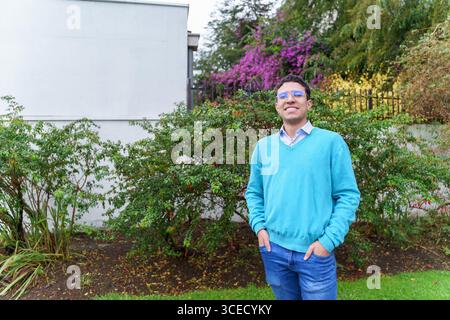 Un jeune homme joyeux de la génération Z avec des lunettes, vêtu d'un pull bleu et d'un Jean, sourit en toute confiance à l'extérieur, regardant la caméra. Le jardin dynamique setti Banque D'Images