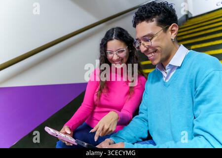 Une femme joyeuse et un homme assis sur un escalier, souriant tout en interagissant avec une tablette. Ils représentent un style de vie moderne de la génération Z, engageant avec DIG Banque D'Images