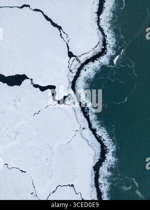La vue aérienne capture la côte hivernale sauvage islandaise avec des terres enneigées rencontrant l'océan bleu glacial. Le contraste saisissant met en évidence la télécommande Banque D'Images