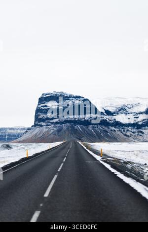 La route asphaltée enneigée s'étend au loin avec des montagnes majestueuses en toile de fond, capturant la beauté lointaine de l'hiver islandais en se Banque D'Images