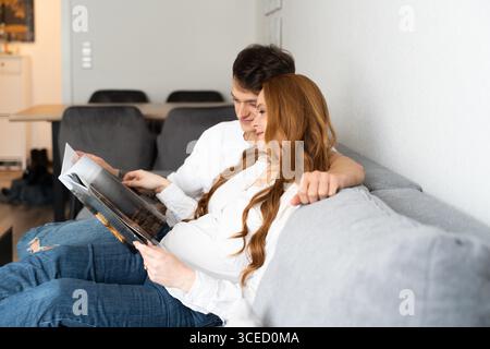 Un jeune couple est assis ensemble sur un canapé confortable, embrassant et lisant un magazine. La future mère repose contre son partenaire, transmettant chaleur et an Banque D'Images