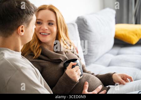 Un couple gai enceinte s'assoit confortablement sur un canapé moderne, partageant un moment chaleureux de connexion. La femme tient une tasse de café en le regardant Banque D'Images