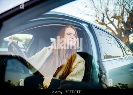 Une belle femme enceinte aux cheveux roux est confortablement assise dans une voiture, regardant dehors avec une expression paisible. Lumière naturelle et cadre extérieur Banque D'Images