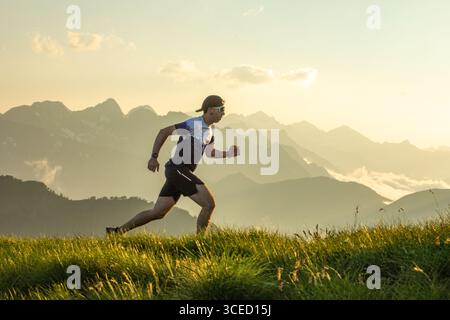 Homme en équipement athlétique courir sur un sentier en herbe avec toile de fond pittoresque de montagne. Le soleil se couche derrière les sommets, projetant une lueur chaude. Porter un chapeau et du soleil Banque D'Images