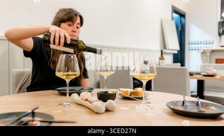 Une femme verse du vin blanc dans des verres sur une table de restaurant pour le dîner. La scène comprend des assiettes avec des hors-d'œuvre, du pain et une table soigneusement agencée Banque D'Images