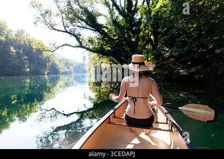 Vue arrière d'une touriste en bikini pagayant un canoë à travers la gorge pittoresque de la rivière Idrijca entourée de falaises et d'arbres, Slovénie. Banque D'Images